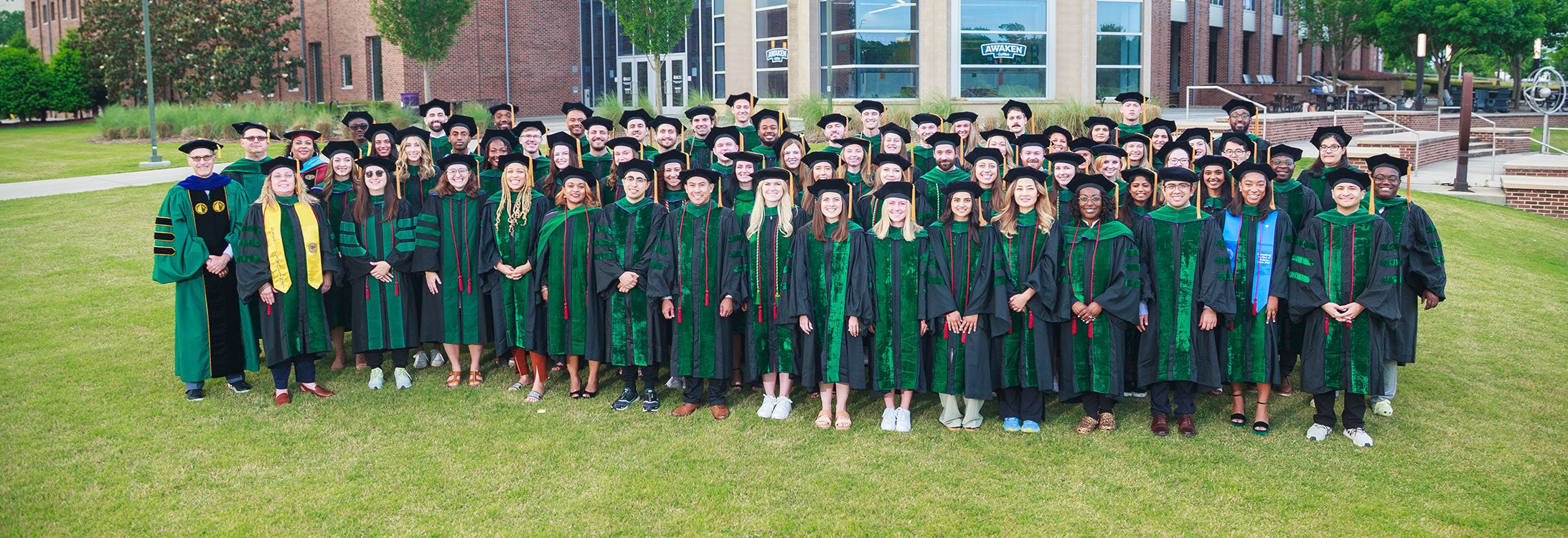A large group of medical school graduates posed together on a grassy lawn in front of a brick campus building. The graduates are wearing traditional black caps and green doctoral robes with colored hoods and stoles indicating their medical specialties. They are arranged in several rows for a formal graduation photo.