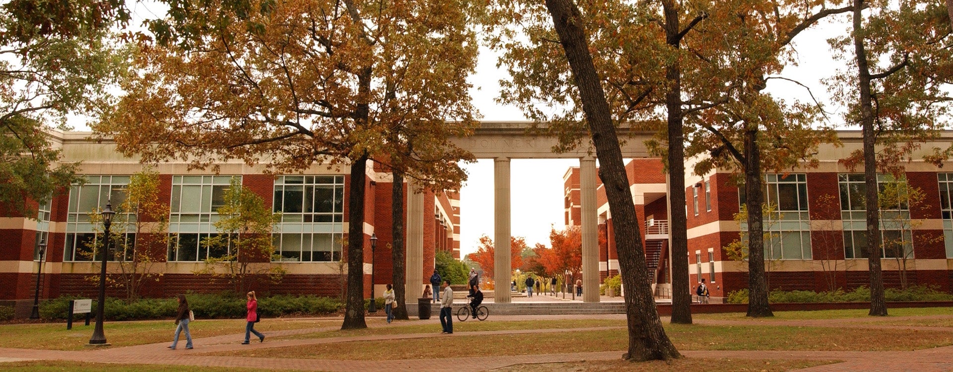 It is a fall day. Students are walking along a tree-lined path in front of buildings on ECU campus.