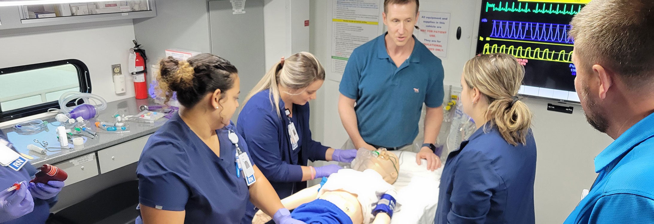 Five medical professionals surround a training dummy on a hospital bed.