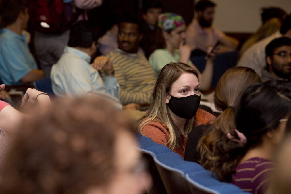 Closeup of a female with long hair wearing a black face mask sitting in a group of people in an auditorium.