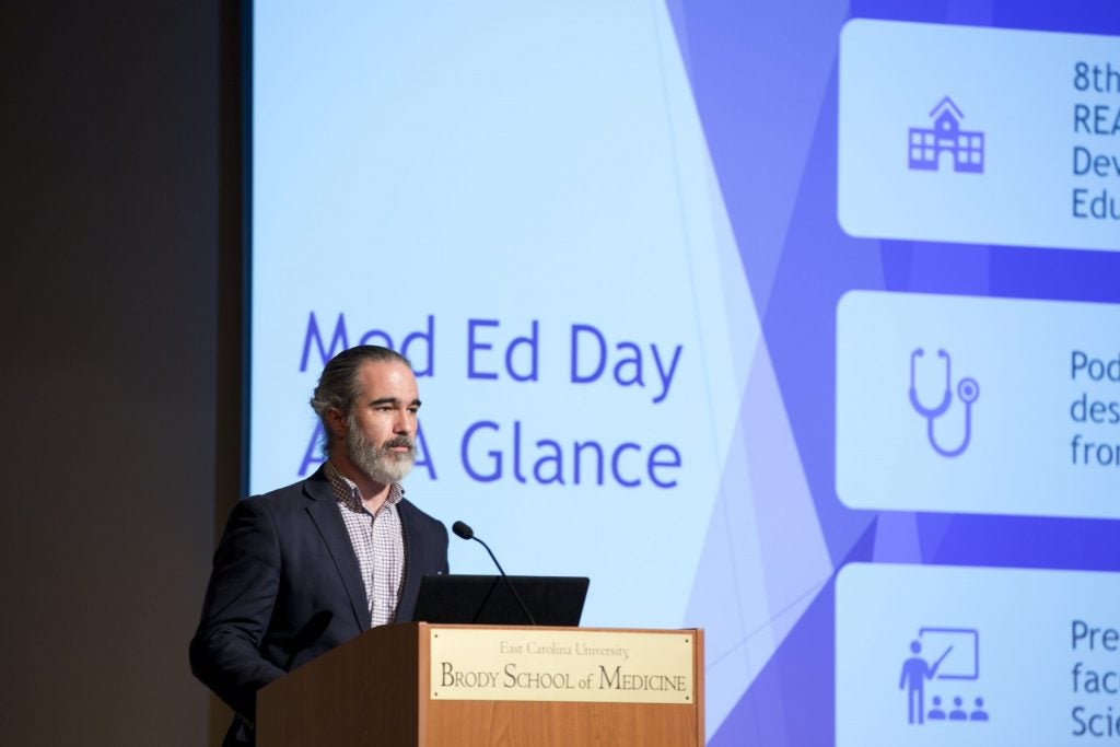 A man with gray facial hair stands behind a podium with a presentation screen behind him that reads: Med Ed Day At A Glance