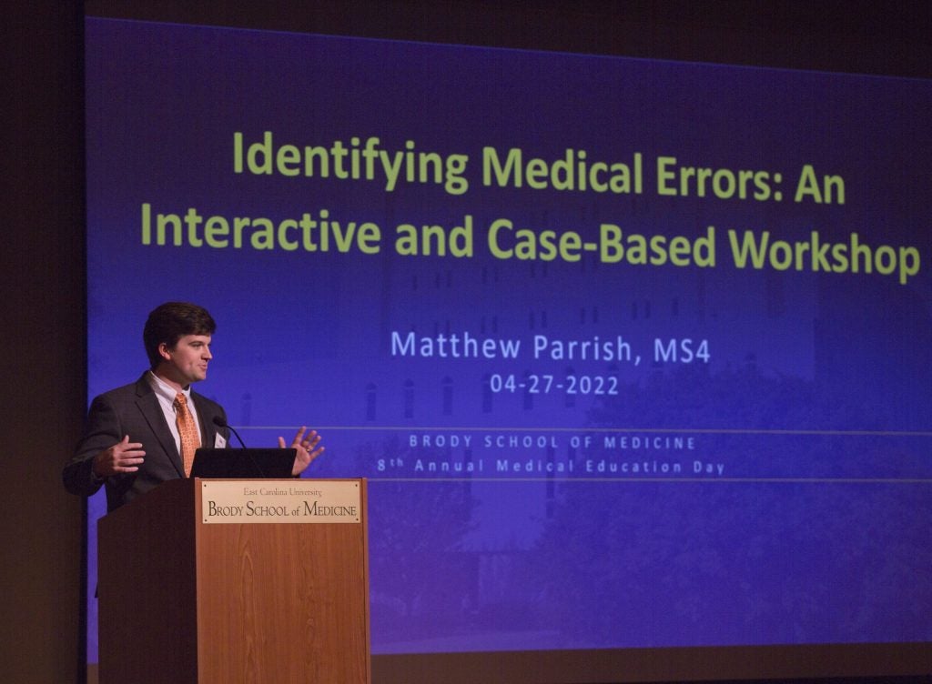 Matthew Parrish stands behind a podium in front of a presentation screen that reads: Identifying Medical Errors: An Interactive and Case-Based Workshop