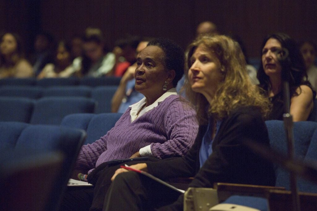 Closeup of two women sitting in an auditorium in a crowd of diverse people.