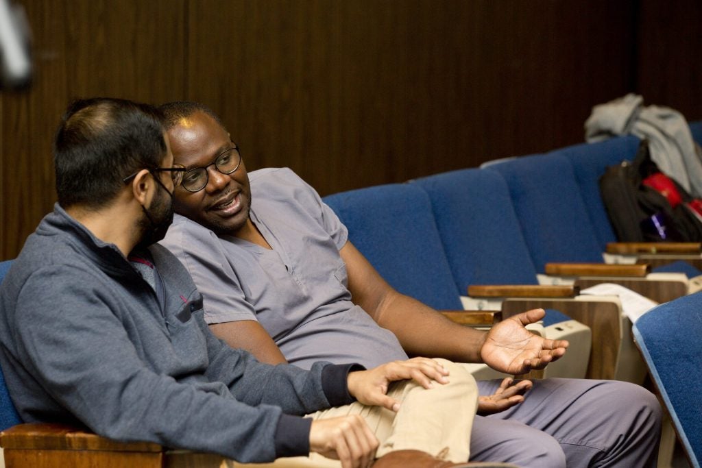 Two men having a conversation in an auditorium. One is wearing scrubs.