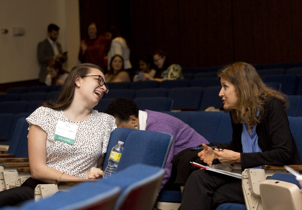 Two women having a conversation in an auditorium. One wears a speaker badge and is laughing.