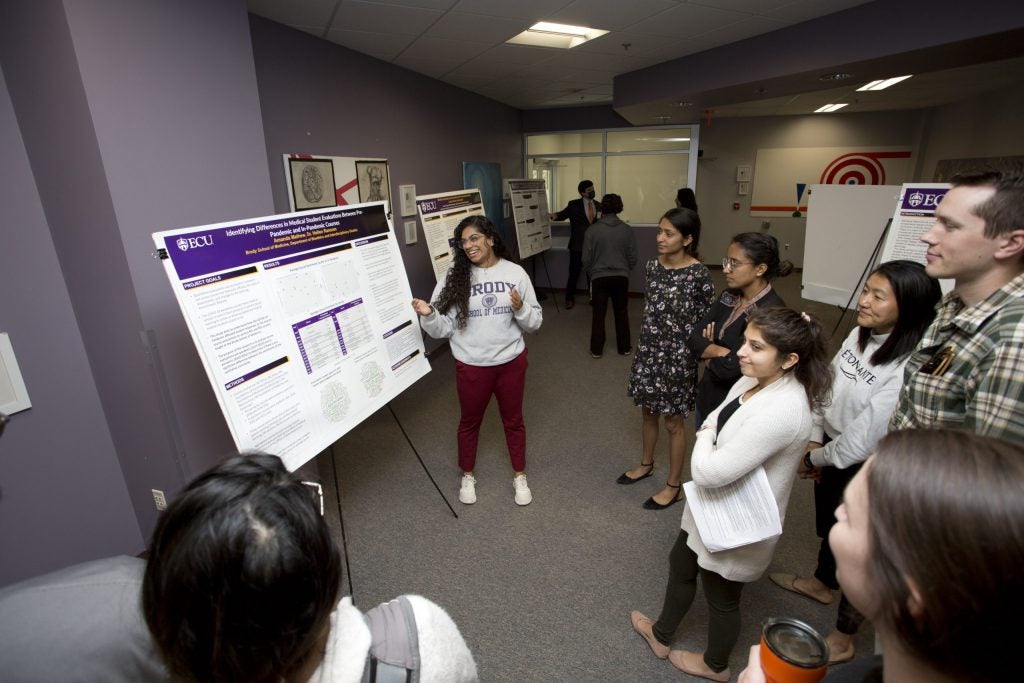 A woman in a Brody School of Medicine sweatshirt standing in front of a poster and speaking to a group of people.