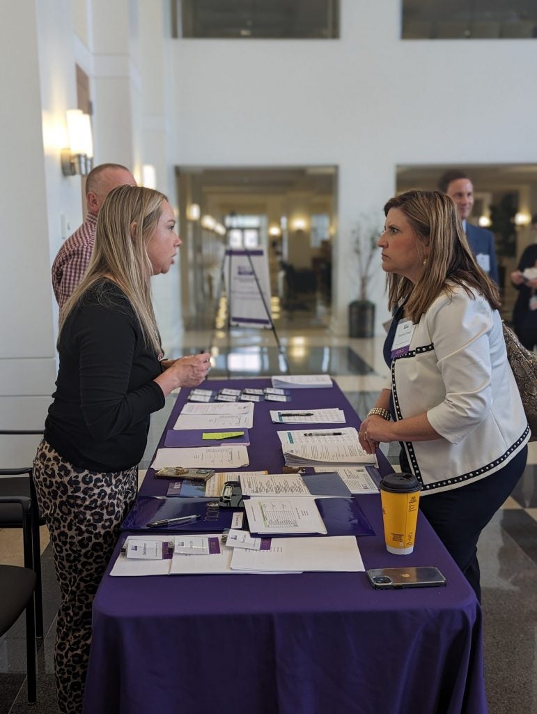 Two women standing with a table between them. A purple tablecloth, name tags, and stacks of paper are on the table.