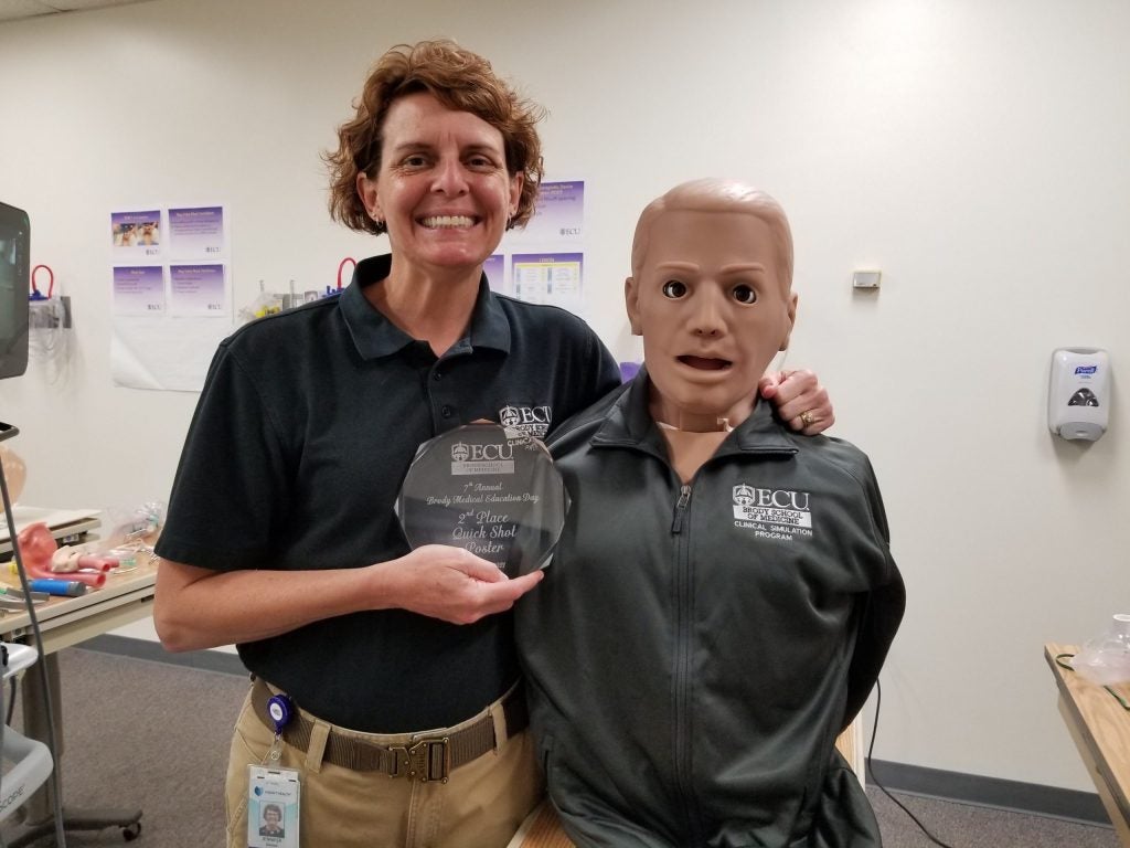 A woman holding a glass second place quick shot poster award standing next to a medical simulation dummy.