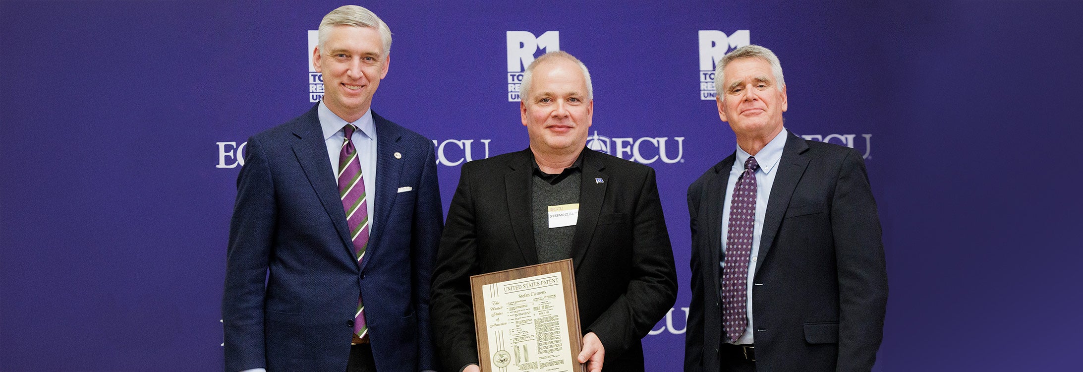 Three men pose for a picture with one man receiving a plaque.